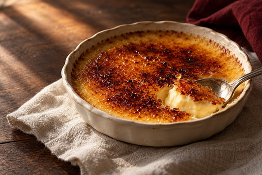 Leite-creme com crosta de açúcar caramelizado, quebrada por uma colher, sobre uma mesa de madeira.